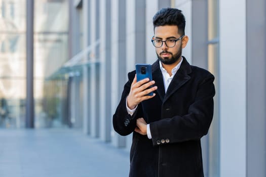 Handsome Indian businessman using smartphone typing text answering messages chatting online looking mobile screen app outdoors. Arabian Hindu freelancer guy in downtown city street. Business people