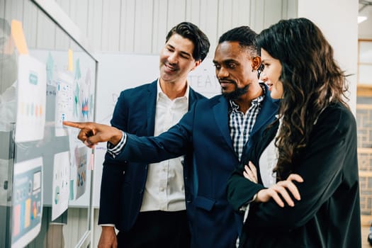 Business professionals in an office meeting use post-it notes on a glass board to brainstorm. Their diverse group emphasizes teamwork, leadership, and successful strategy discussions.