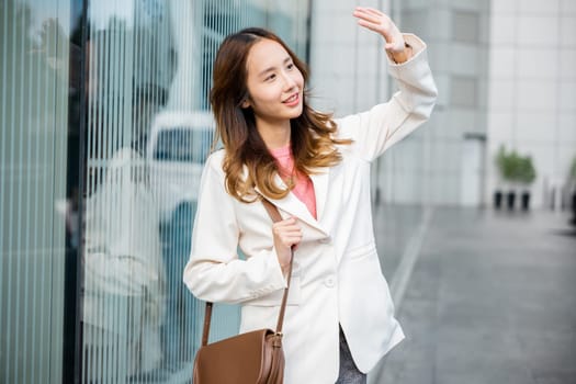 Portrait of confidence young businesswoman standing outside office building in city raise your hand to shade the sun. Happy woman wearing white suit jacket with brown bag at sunlight outdoors.