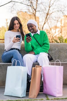 two young female friends using mobile phone sitting in a city park with shopping bags, concept of technology of communication and urban lifestyle