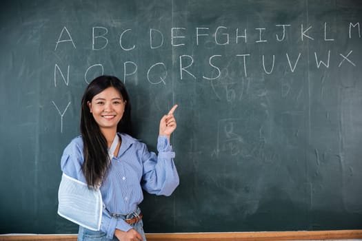 A woman is pointing at the alphabet on a blackboard. Arm broken. Teacher confident broken arm after accident and wear arm splint for treatment, sling support hand