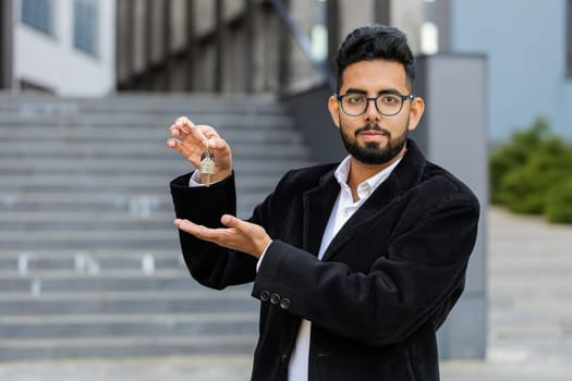 Indian businessman real estate agent lifting hand showing keys of new home house apartment buying or renting property, mortgage loan, investment outdoors. Arabian Hindu freelancer guy in downtown city