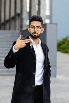 Indian businessman real estate agent lifting hand showing keys of new home house apartment buying or renting property, mortgage loan, investment outdoors. Arabian Hindu freelancer guy in downtown city