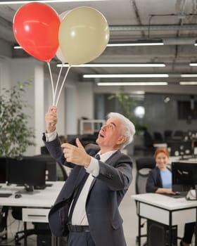 Portrait of a cheerful mature business man holding balloons in the office. Vertical photo