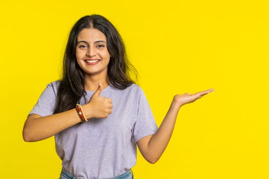 Indian woman showing thumbs up and pointing right empty place, advertising area for commercial text copy space for goods promotion advertisement. Hindu girl isolated on yellow background. Lifestyles