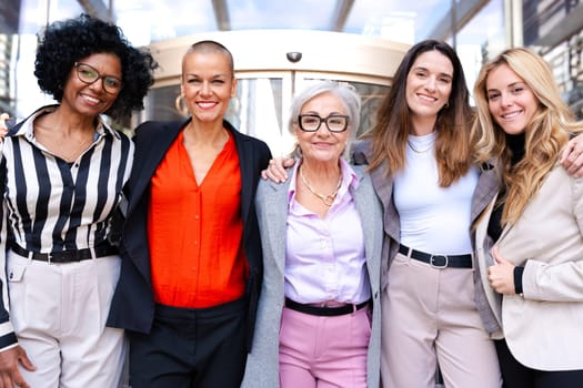 Five businesswomen standing, side by side, holding each other by the waist looking at the camera, happy. Suitable for team, friendship and diversity concepts.