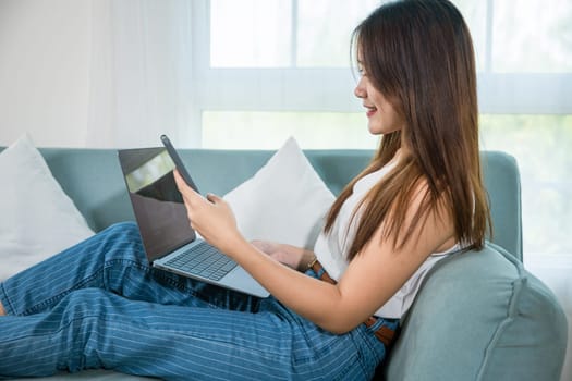 Happy woman use notebook browsing website and payment shopping online with smartphone, Asian young female using smart mobile phone while sitting on sofa in living room at home with laptop computer