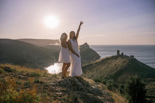 Two young girls are standing on a hillside, one of them wearing a white dress. The sun is shining brightly, creating a warm and inviting atmosphere. The girls seem to be enjoying their time together