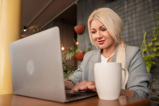 A woman is sitting at a table with a laptop and a white coffee cup. She is smiling and she is enjoying her time