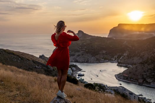 Happy woman standing with her back on the sunset in nature in summer with open hands posing with mountains on sunset, silhouette. Woman in the mountains red dress, eco friendly, summer landscape active rest.