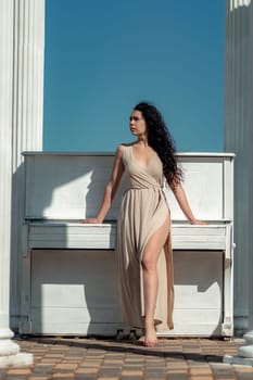 A woman in a long dress stands in front of a piano. The piano is white and has a few keys visible. The woman is posing for a photo, and the overall mood of the image is calm and serene