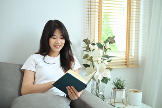 Happy young asian woman in casual clothes reading book on couch, spending free time at home.