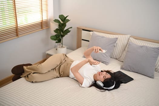 Relaxed young woman listening to music with headphones and using smartphone while lying in bed.