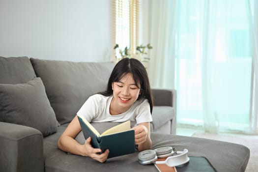 Carefree young asian woman in casual clothes lying on couch and reading book.