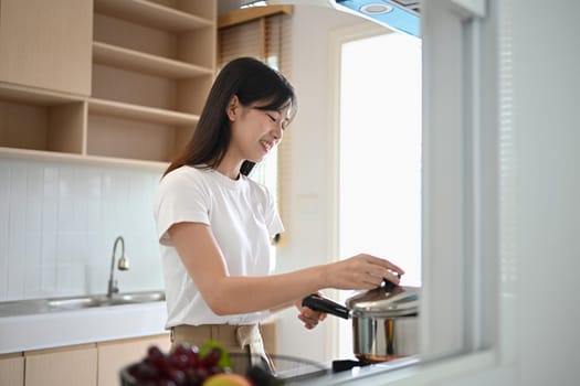 Young beautiful woman standing by the stove, preparing lunch in modern kitchen.