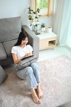 Happy young woman using digital tablet sitting carpet in living room.