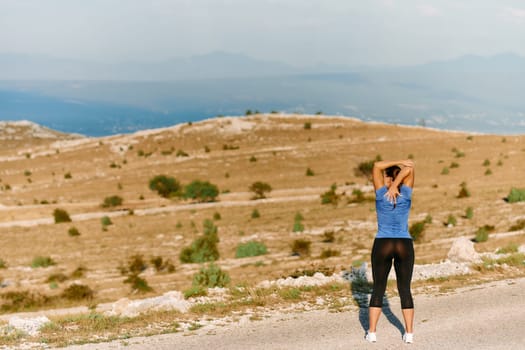 A determined female athlete stretches her muscles after a strenuous run through rugged mountain terrain, surrounded by breathtaking rocky landscapes.