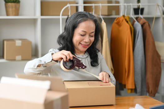 Smiling senior woman small business owner packing parcel boxes of product for shipping to customer.