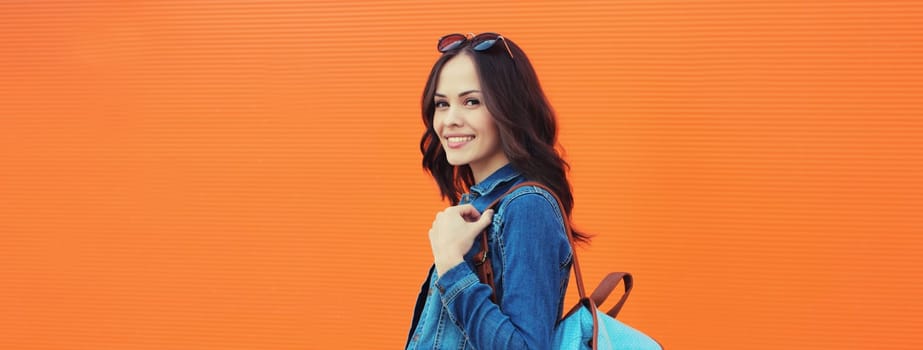 Summer portrait of happy smiling brunette young woman with backpack walking along city street, orange background