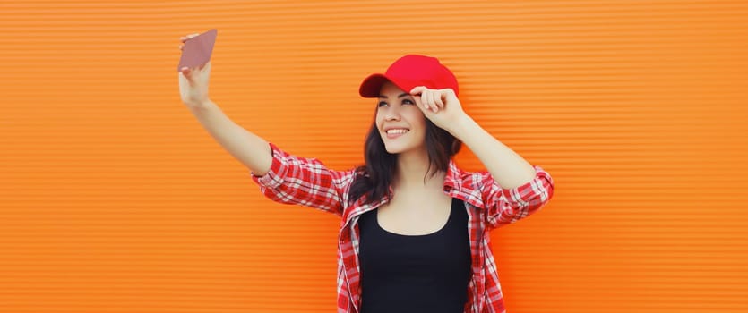 Summer portrait of happy cheerful smiling young woman taking selfie with mobile phone in red baseball cap on colorful orange background on city street