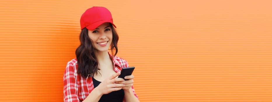 Portrait of happy smiling young woman with mobile phone in summer red baseball cap on colorful orange background, blank copy space for advertising text