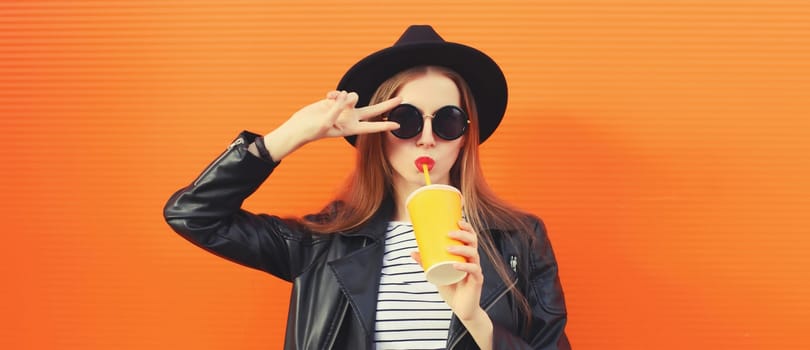 Portrait of stylish young woman drinking fresh juice posing in black rock style leather jacket, round hat on orange background
