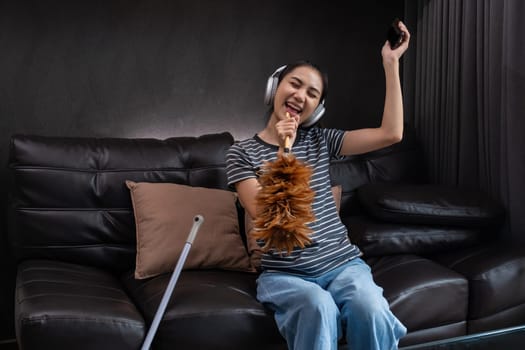 Smiling woman dusting with feather duster and listening to music on headphones. Concept of multitasking and joyful housework.