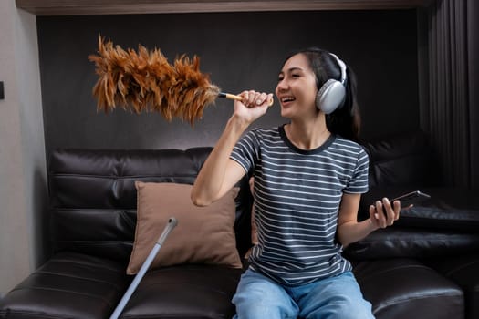 Smiling woman dusting with feather duster and listening to music on headphones. Concept of multitasking and joyful housework.