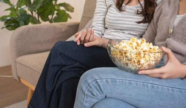 Close-up of two women holding hands with bowl of popcorn on couch. Concept of friendship and comfort.