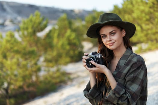 Stylish woman in plaid shirt and hat capturing memories with vintage camera in outdoor setting