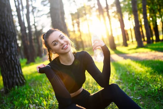 A smiling young woman in a sporty black suit holds a water bottle in the woods