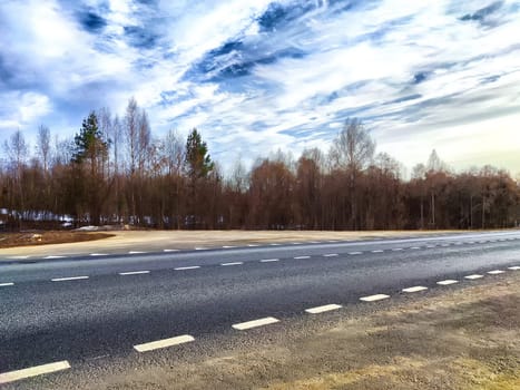 A lonely country road stretches alongside barren trees under a clear, partly cloudy sky on peaceful winter afternoon