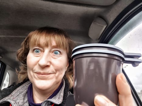 Funny Middle aged woman with a coffee mug in car taking selfie while enjoying drive. Female mature driver posing inside car. Funny happy tourist girl