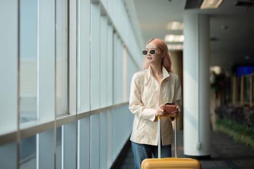 Asian woman with sunglasses holding luggage in airport terminal. Concept of travel, style, and anticipation.