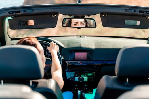 A woman is driving a convertible car with the top down. She is looking at her reflection in the rearview mirror