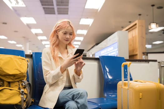Asian woman using smartphone in airport with luggage. Concept of travel, technology, and waiting.