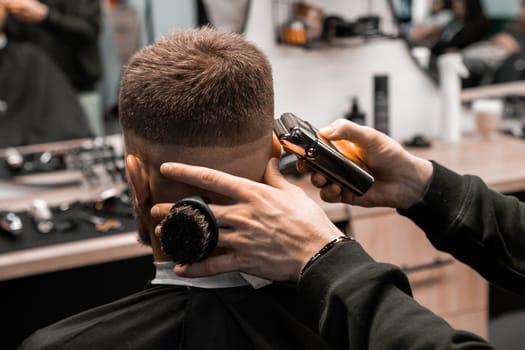 Hairstylist removes hair behind client ear and occiput with shaver in barbershop closeup. Barber uses modern machine for stylish haircut in salon