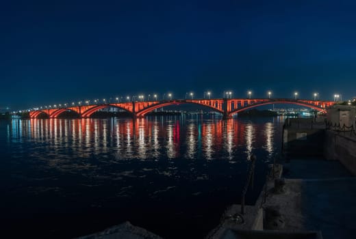 A bridge crossing over a body of water during the nighttime, set against a cityscape with the sky and horizon visible in the background