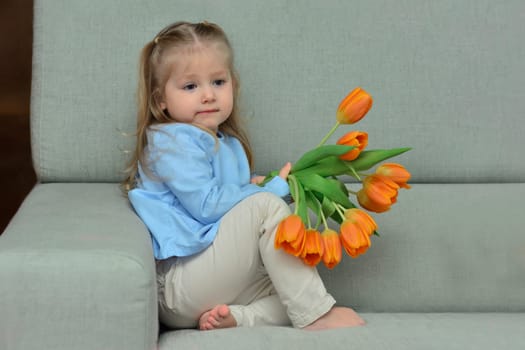 A three-year-old girl sits on the sofa and holds orange tulips in her hands in a room