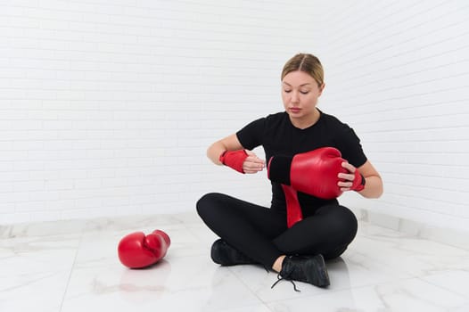 Portrait of European young woman boxer with boxing gloves, ready for match, sitting against white wall background. Sport, cardio workout, boxing training or endurance for energy, strong or power.