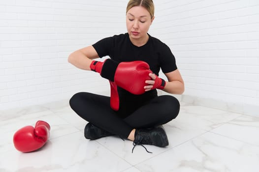 Young blonde female boxer fighter preparing for boxing training, putting on red boxing gloves, sitting in lotus pose against white brick background. Athletic discipline, sport, combat, martial art.