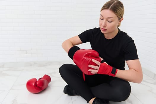 Portrait of European woman boxer putting on red boxing gloves, sitting against white wall background. The concept of athletic discipline, sport, combat, martial art.