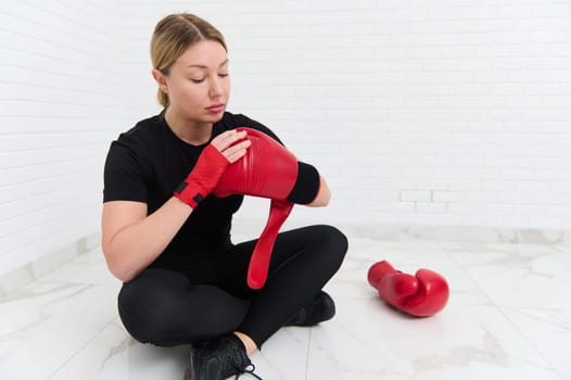 Young Caucasian woman boxer fighter putting on red boxing gloves, sitting against white background. The concept of athletic discipline, sport, combat, martial art.