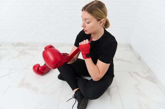 View from above of a young European woman boxer fighter putting on red boxing gloves, sitting against white wall background. The concept of athletic discipline, sport, combat, martial art.