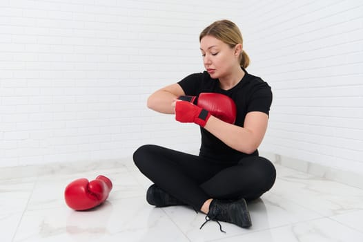 Full size shot of a young Caucasian woman boxer fighter putting on red boxing gloves, sitting against white wall background. The concept of athletic discipline, sport, combat, martial art.