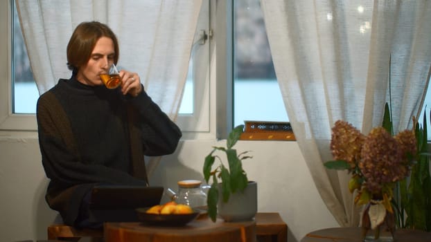 Young man drinks tea and looks at tablet in cafe. Media. Handsome young man is drinking tea alone and looking at tablet. Student is relaxing with tablet in cozy cafe.