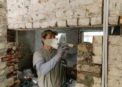 One young Caucasian man lays bricks on fresh cement in a doorway by tapping with the handle of a spatula and standing on a stepladder to the right in an old house, close-up side view.