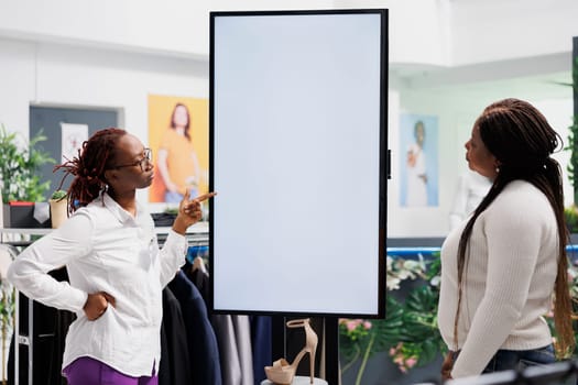 Shopping mall worker explaining shoes brand features to client using blank smart display. African american store assistant showcasing new collection to buyer on digital empty screen in boutique