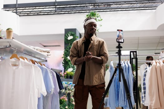 African american blogger recording promotional video in clothing store to attract customers. Stylist talking about new fashion collection on smartphone camera and advertising brand in shop
