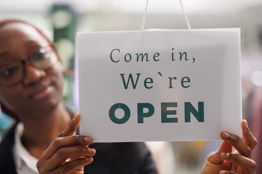 Fashion clothes shop manager holding opening signboard with come in message. Apparel store young african american woman assistant hanging sign informing clients about workday beginning